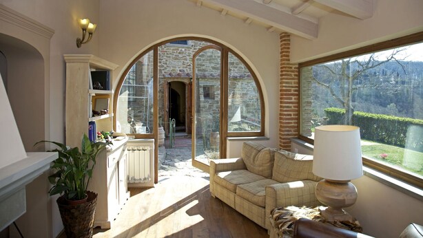 Ein gemütliches Zimmer mit einem großen Bogenfenster, einer Couch, einer Lampe und Blick auf ein Steinhaus.