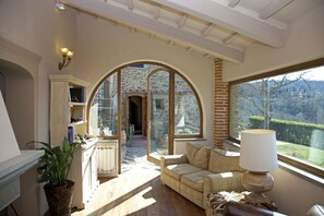 Una habitación acogedora con un gran ventanal de arco, un sofá, una lámpara y vista a un edificio de piedra.