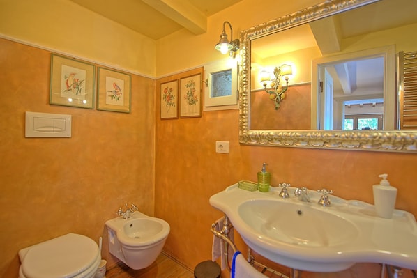 Une salle de bain équipée d’un bidet, de toilettes et d’un lavabo. Des tableaux encadrés ornent le mur, et un miroir est placé au-dessus du lavabo.