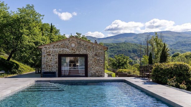 Un edificio de piedra con piscina, rodeado de vegetación y montañas.