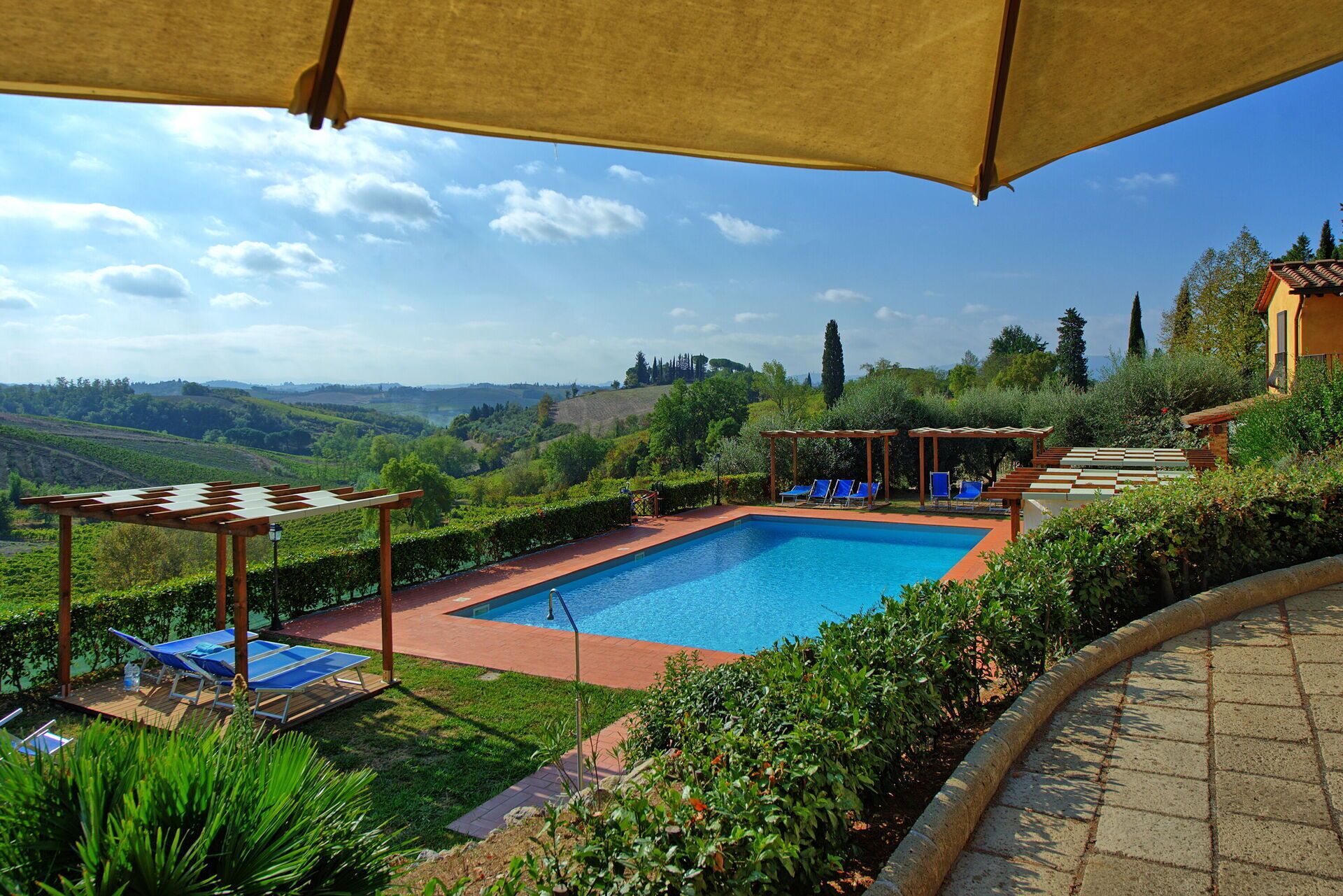 A rectangular swimming pool with a pergola and lounge chairs, surrounded by lush greenery and a scenic hillside view.