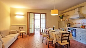 A dining area with a table set for four, a sofa, a kitchen with a stove, and a large window with curtains. - Casa Rossa 4 - Apartment with swimming pool (Gorgognano)