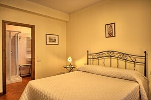 A bedroom with a bed, a nightstand with a lamp, a framed picture on the wall, and a bathroom visible through an open door. - Casa Rossa 4 by PosarelliVillas (Gorgognano)