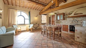 Eine gemütliche Küche mit Fliesenboden, Holzbalken und einem Steinofen. Es gibt einen Esstisch mit Stühlen und ein Sofa im Wohnbereich.