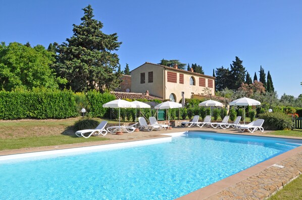 A swimming pool with lounge chairs and umbrellas, surrounded by greenery and a building with a tiled roof.