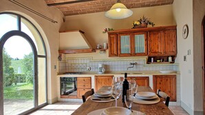 Un coin repas avec une table en bois dressée pour le repas, une cuisine avec des placards en bois et une vue sur un jardin par le biais d’une grande fenêtre.