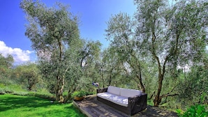 A wooden deck with a dark wicker sofa and cushions, surrounded by lush greenery and trees.