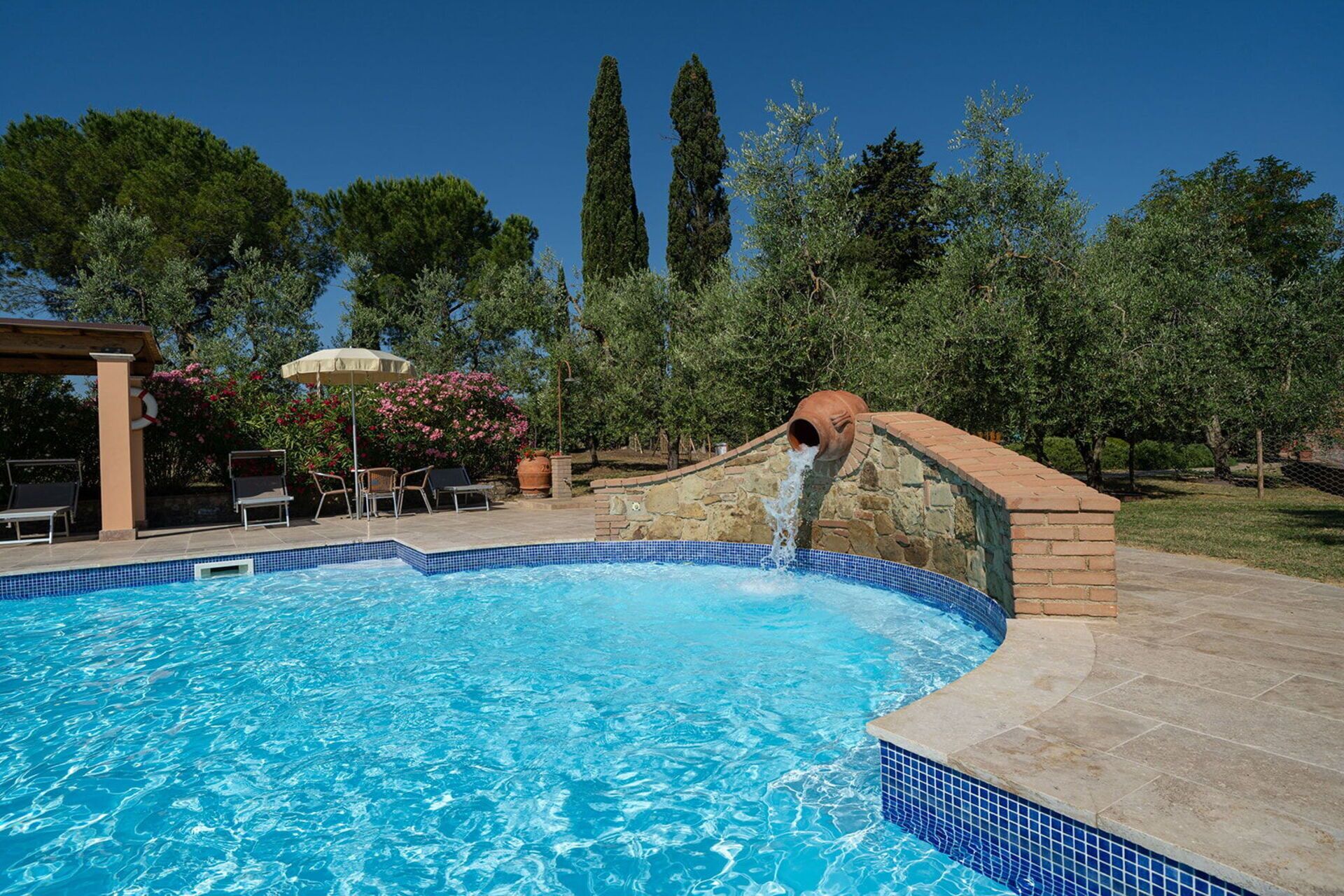 A swimming pool with a water feature, surrounded by a patio area with chairs and a pergola.
