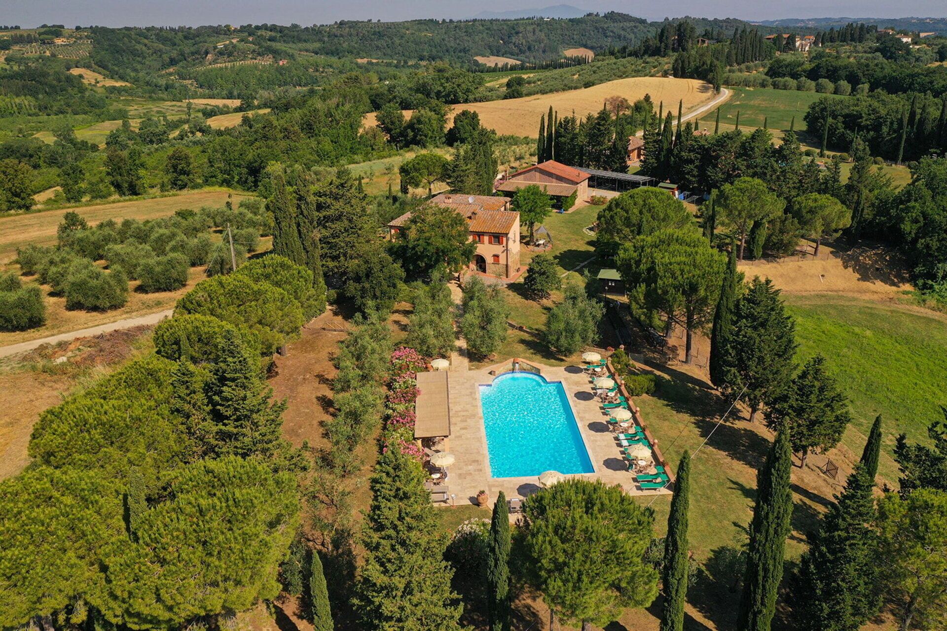 Vue aérienne d’une propriété dotée d’une piscine, entourée de chaises longues et de parasols, située dans un paysage verdoyant et luxuriant, avec des collines douces en arrière-plan.
