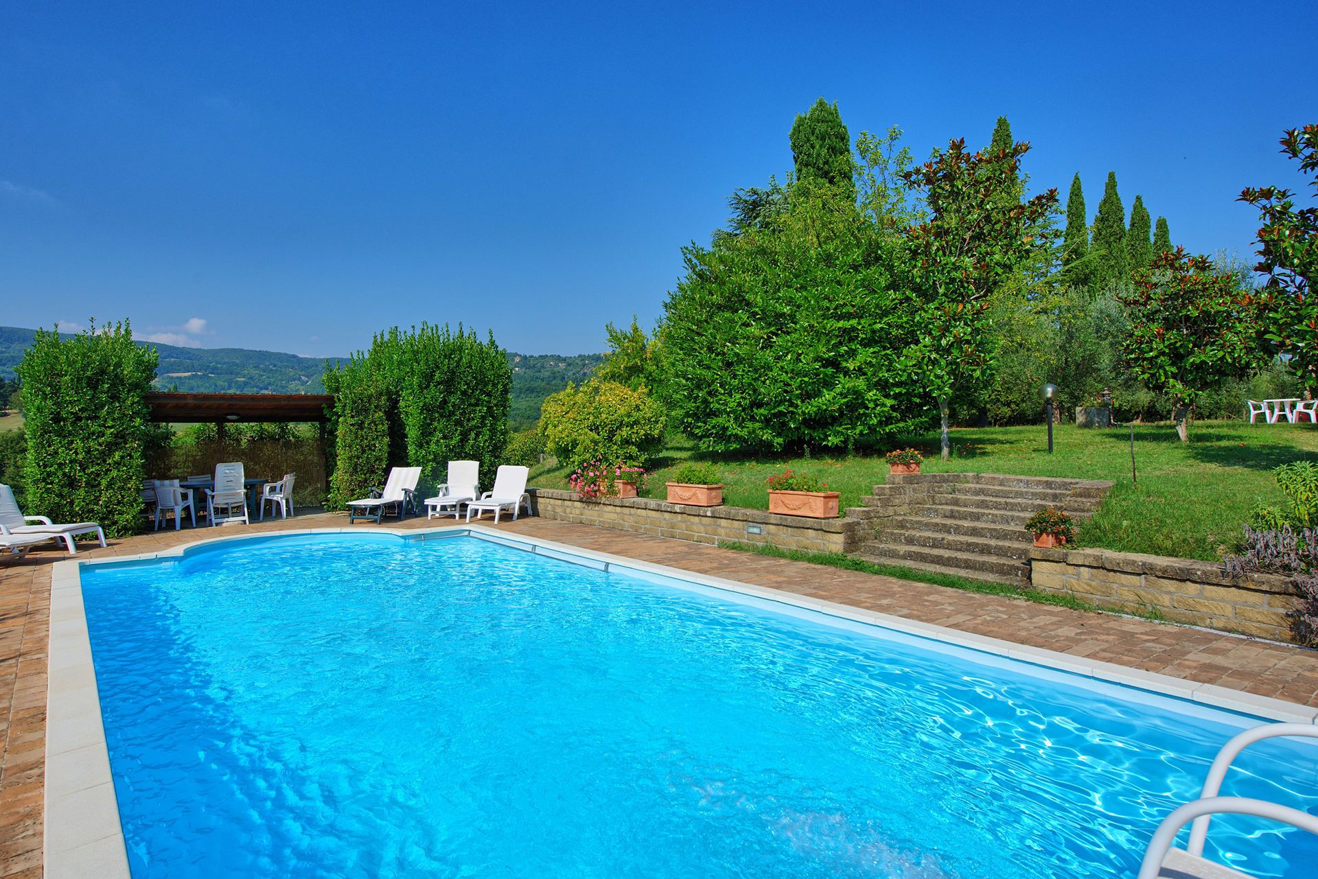 A swimming pool with lounge chairs and a pergola, surrounded by greenery and a view of hills.