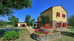 Une maison à deux étages, avec des fondations en pierre et des volets rouges, entourée d’un jardin bien entretenu, agrémenté de plantes fleuries et d’un espace détente avec des chaises.