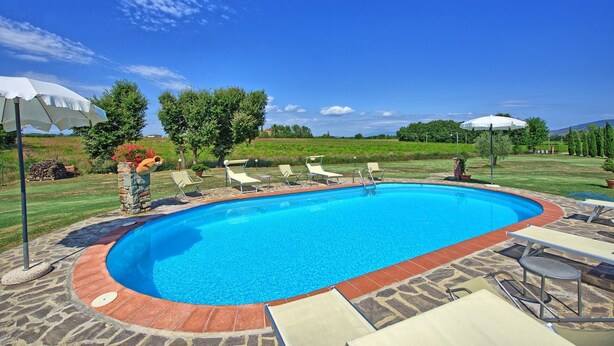 Une piscine entourée d’une terrasse en pierre et de verdure, avec des chaises longues et des parasols.
