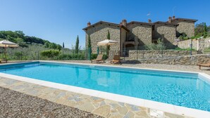 A swimming pool with stone tiles, two stone buildings with chimneys, and a clear blue sky.