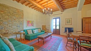 A living room with a stone wall, a green sofa, a wooden coffee table, and a dining area with a table and chairs. - Il Frantoio - Villa with private pool (Montelupo Fiorentino)