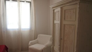 Ein Zimmer mit weißem Ohrensessel, einem großen Fenster mit transparenten Vorhängen und einem weißen Schrank mit dekorativen Verzierungen.