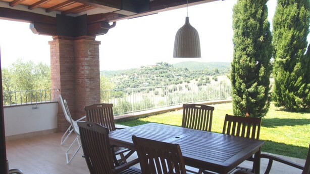 Eine überdachte Terrasse mit Holztisch und -stühlen, einer hängenden Pendelleuchte und Blick auf eine bewaldete Hügellandschaft.