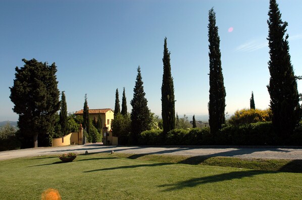 A landscaped garden with a yellow building, tall cypress trees, and a clear sky.