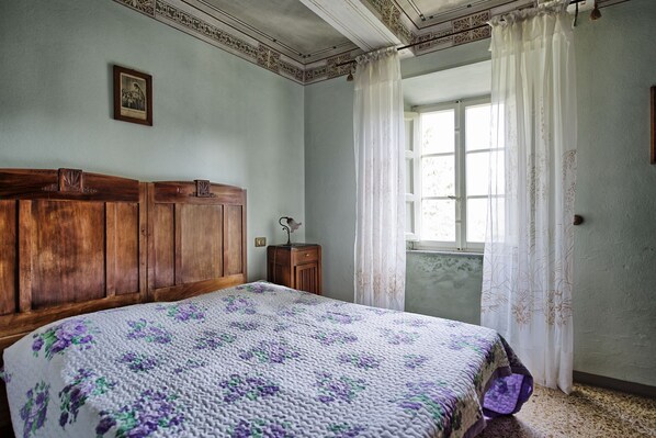 A bedroom with a wooden headboard, a bed with a floral quilt, a nightstand, a window with sheer curtains, and a framed picture on the wall.