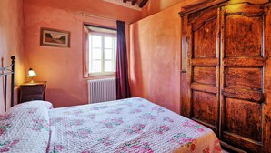 Quarto com guarda-roupa de madeira, cama com colcha florida, radiador, janela com cortinas e luminária na parede.
