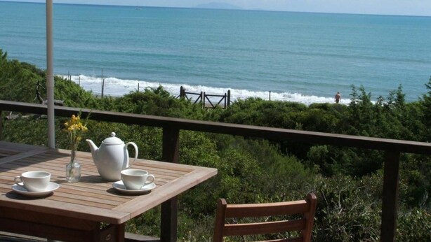 A wooden table with a white teapot and cups, an umbrella, and a view of the ocean.