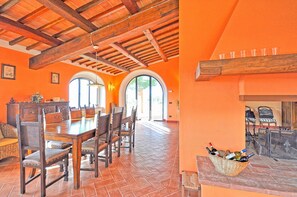 A dining area with a wooden table and chairs, a fireplace with a basket of wine bottles, and a view of the outdoors through arched windows.