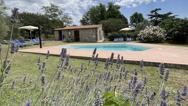 Un bâtiment en pierre avec un toit recouvert de tuiles rouges, une piscine et plusieurs chaises longues bleues.