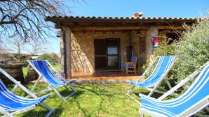 Une maison en pierre avec un toit de tuiles, une porte en bois et un petit porche. On y trouve deux fauteuils de jardin à rayures, posés sur la pelouse devant la maison.