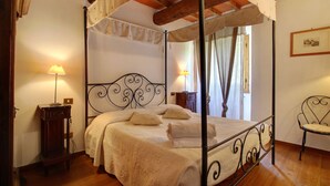 Una cama con dosel sobre cuatro columnas, suelo de madera, dos mesitas de noche con lámparas y una silla.