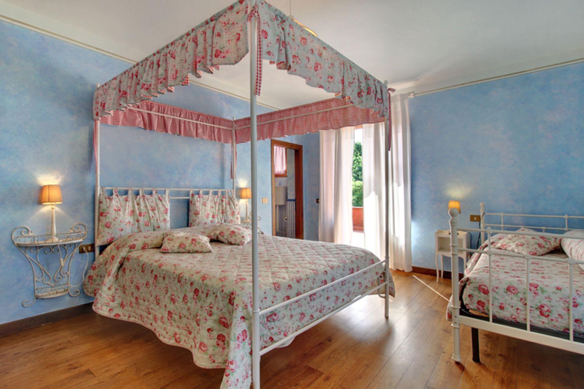 A bedroom with a four-poster canopy bed, a smaller single bed, wooden flooring, and a view of the outdoors through a window.