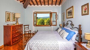 Una camera da letto con soffitto in legno, un letto con copriletto decorato, un comò, una sedia e una finestra vista verde.