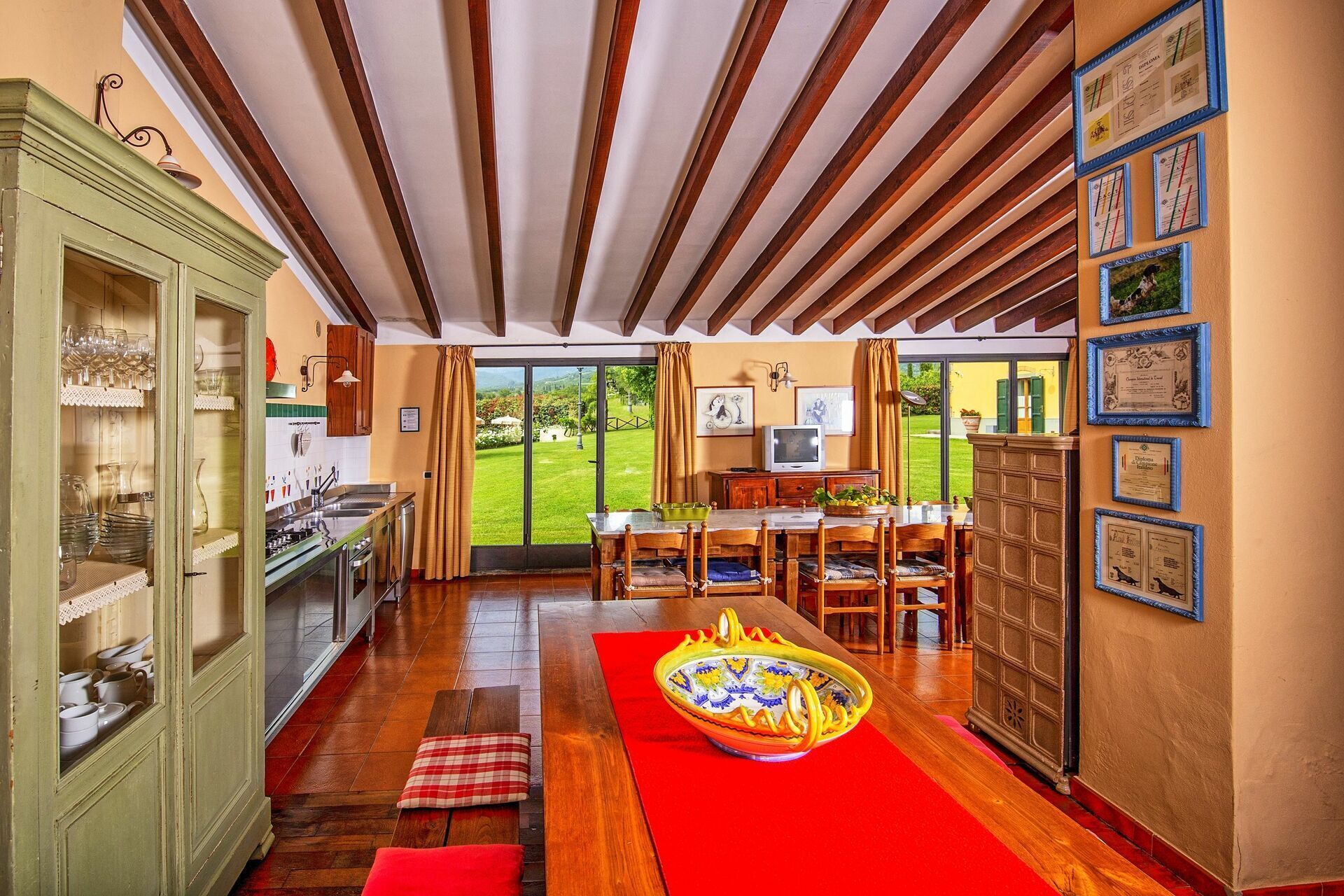 A spacious kitchen with a dining area, wooden beams on the ceiling, and a view of a green garden through the glass door.
