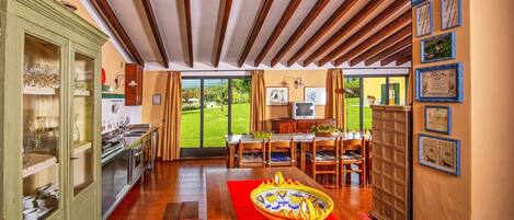 A spacious kitchen with a dining area, wooden beams on the ceiling, and a view of a green garden through the glass door.