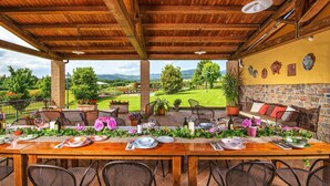 Zona pranzo all'aperto con tavolo in legno apparecchiato per un pasto, circondato da sedie, con vista su un paesaggio verde e montagne sullo sfondo.