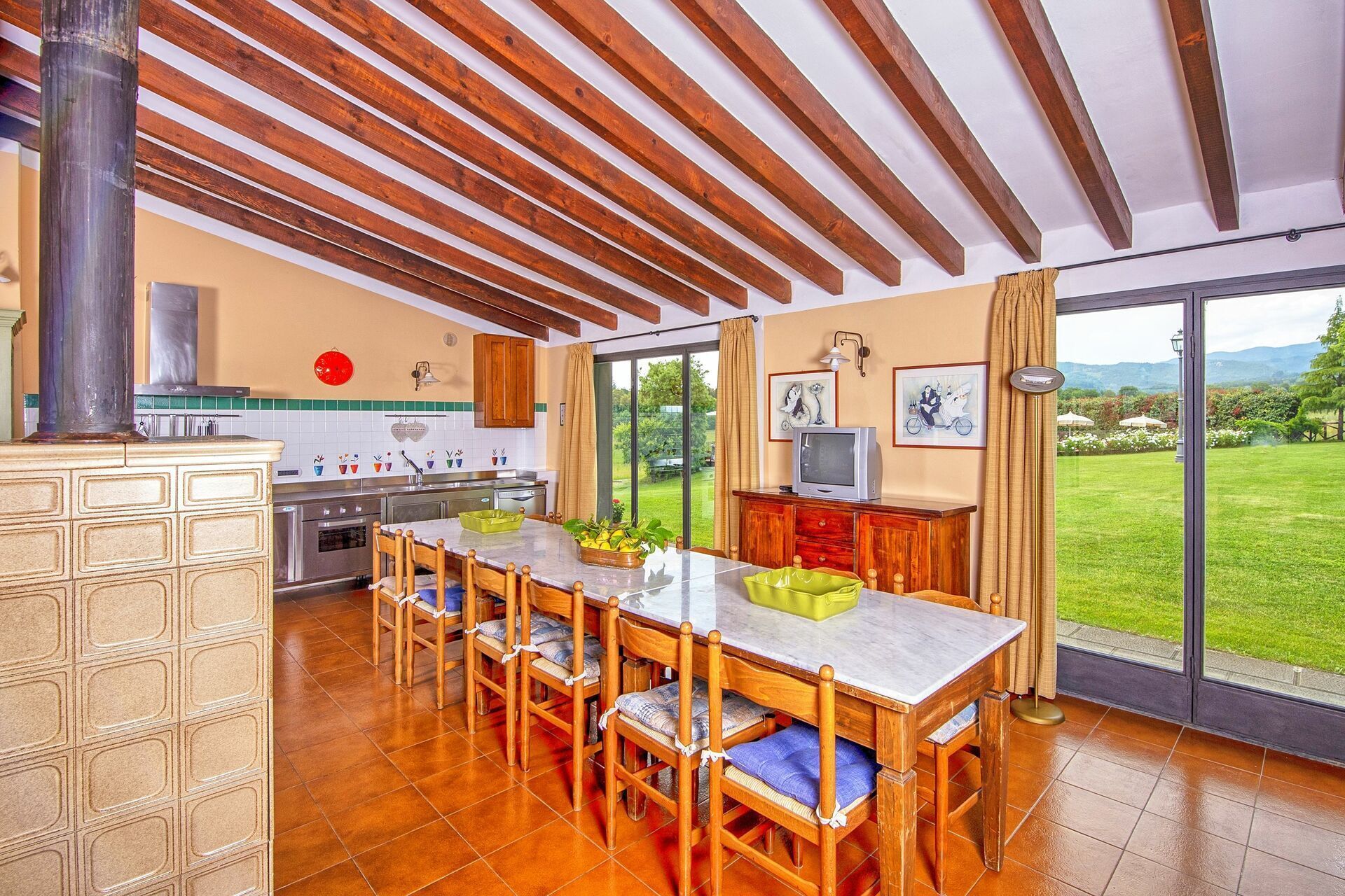 A kitchen with wooden beams, a dining area with chairs, a TV, and a view of a garden.