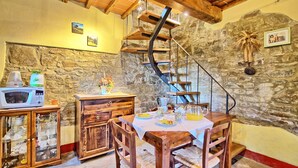 A rustic dining area with a wooden table, chairs, and a stone wall. A spiral staircase is visible in the background.