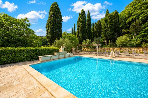 A rectangular swimming pool with clear blue water, surrounded by stone steps and a pergola with chairs.
