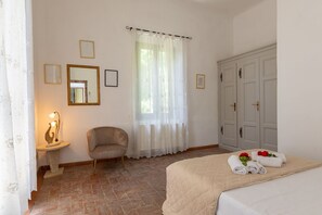 A bedroom with a bed, a chair, a bedside table, a window with curtains, a wardrobe, and framed pictures on the wall.
