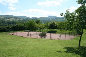 En tennisbana omgiven av frodigt, grönt gräs och träd, med böljande kullar och en klar himmel som bakgrund.
