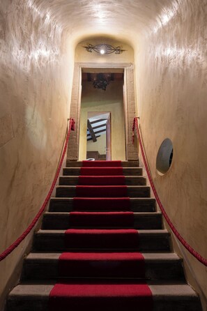 Un escalier dont les marches sont recouvertes d’une moquette rouge, menant à une porte, de part et d’autre de laquelle se trouvent des murs et une barrière faite d’une corde rouge.