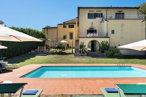 Une piscine avec des chaises longues et des parasols, un bâtiment doté d’un balcon et un jardin bien entretenu.