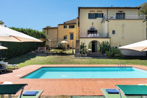 A swimming pool with lounge chairs and umbrellas, a building with a balcony, and a well-maintained garden.
