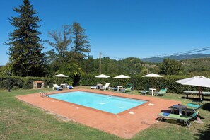 Une piscine rectangulaire bordée de carreaux, entourée de pelouse et d’une terrasse en briques. Plusieurs chaises longues et parasols sont disposés autour de la piscine.