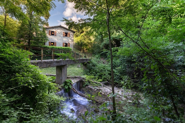 Una casa de piedra con contraventanas de madera, un puente de piedra y una cascada en un bosque exuberante.