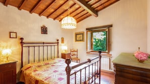 Une chambre à coucher avec un lit en bois, une table de chevet, une commode, une fenêtre donnant sur un paysage verdoyant et un lustre.