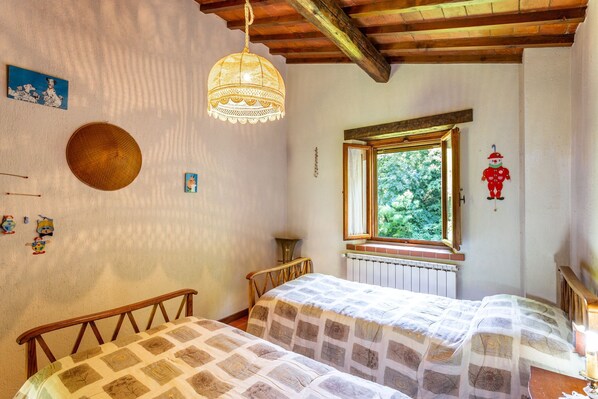 Un dormitorio con dos camas de madera, una lámpara colgante, una ventana con vistas a la vegetación y un radiador.