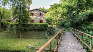 Une maison en pierre avec des volets en bois, un chemin de gravier menant à la maison et un pont en bois enjambant un ruisseau.