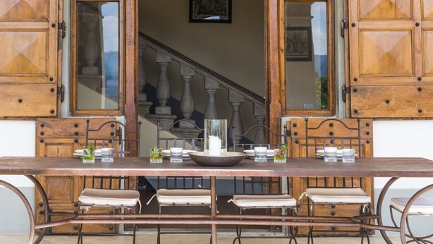 Un área de comedor al aire libre con una mesa y bancos de madera, una escalera al fondo y una lámpara colgante.