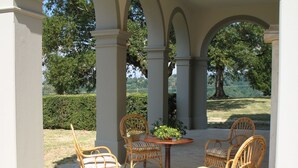 Un patio cubierto con sillas de mimbre y una mesa, rodeado de columnas y vistas a un jardín paisajístico.