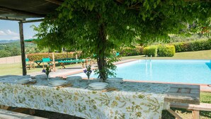 Zona de comedor junto a la piscina con una mesa preparada para cuatro personas, rodeada de vegetación y con vistas a una piscina.