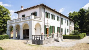 Un edificio de dos pisos con fachada blanca y contraventanas verdes, que presenta un balcón con barandilla y una entrada cubierta con arcos.